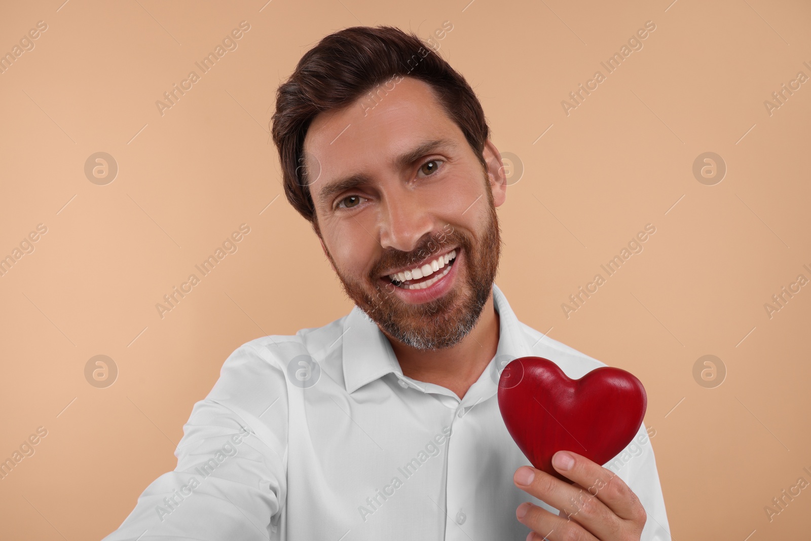 Photo of Happy man holding red heart and taking selfie on beige background