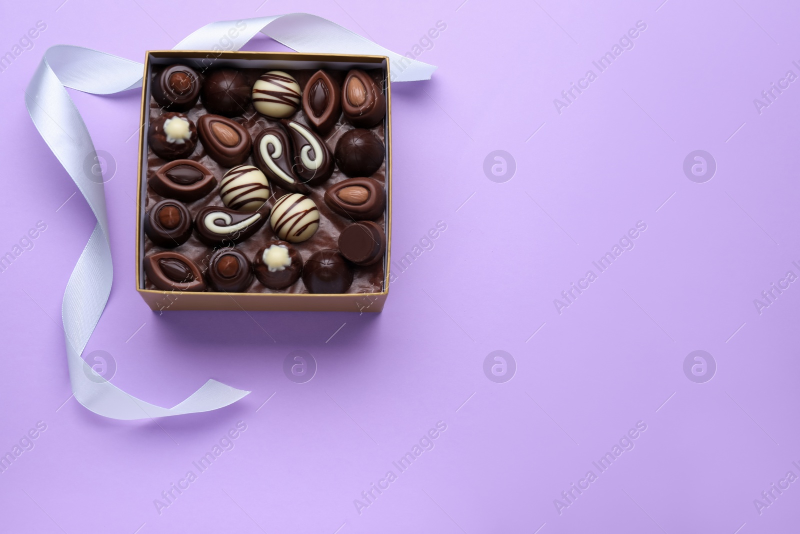 Photo of Box of delicious chocolate candies and color ribbon on violet background, top view. Space for text