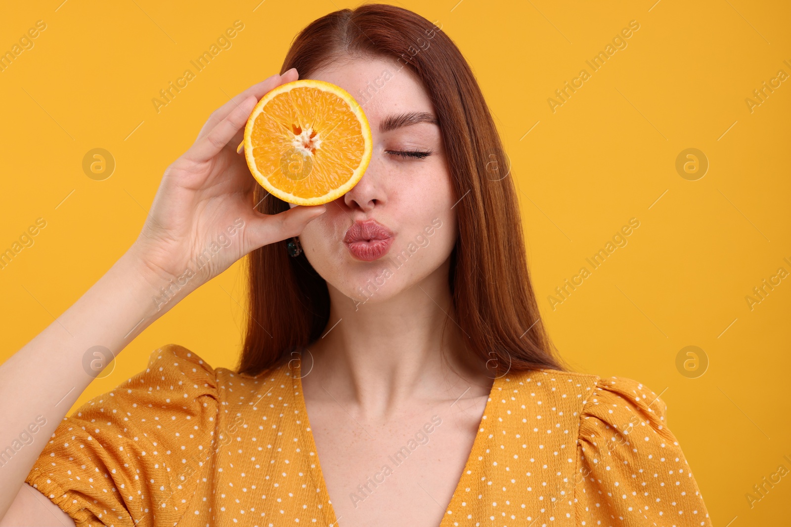 Photo of Beautiful woman covering eye with half of orange and sending air kiss on yellow background