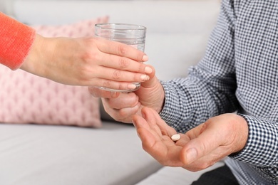 Photo of Woman giving glass of water to senior man with pill indoors, closeup