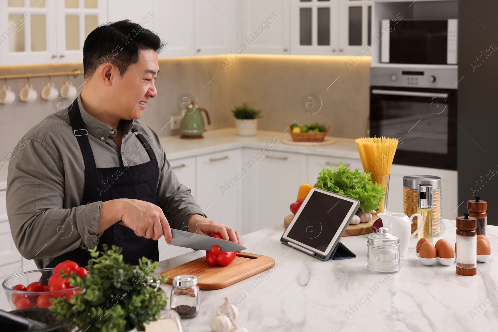 Photo of Cooking process. Man using tablet while cutting fresh bell pepper at countertop in kitchen