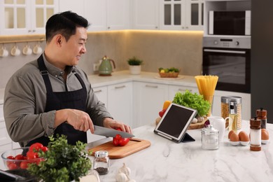 Photo of Cooking process. Man using tablet while cutting fresh bell pepper at countertop in kitchen