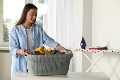 Photo of Young woman with basket full of clean laundry at table indoors