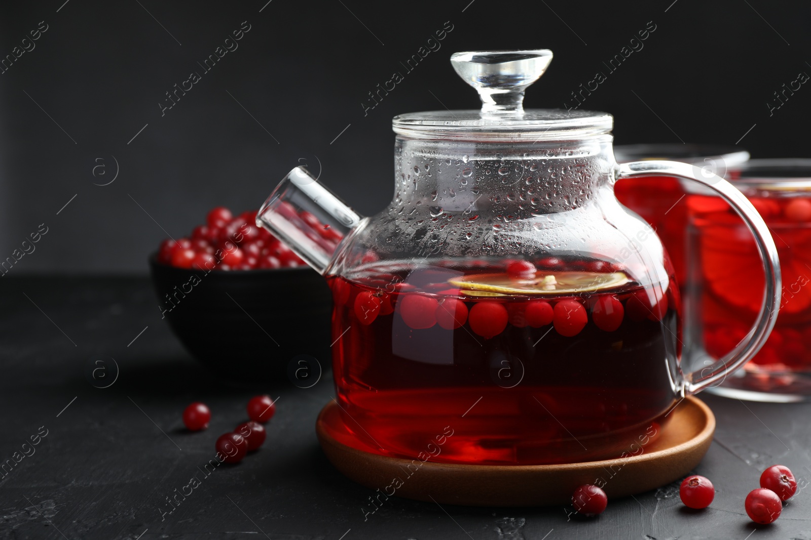 Photo of Tasty hot cranberry tea with lemon in teapot and fresh berries on black textured table