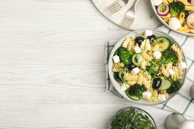 Photo of Bowl of delicious pasta with cucumber, olives, broccoli and cheese on white wooden table, flat lay. Space for text