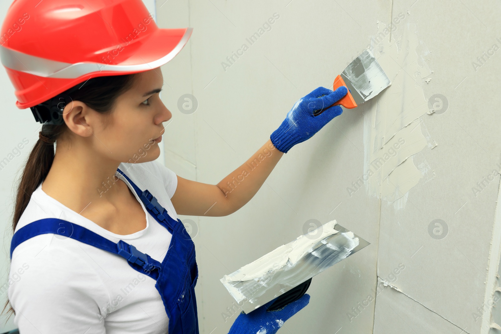 Photo of Professional worker in hard hat plastering wall with putty knives indoors