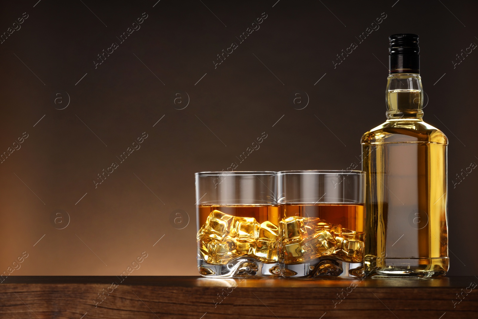 Photo of Whiskey with ice cubes in glasses and bottle on wooden table against brown background, space for text