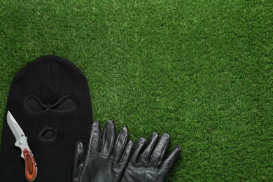 Black knitted balaclava, gloves and knife on green grass, flat lay. Space for text