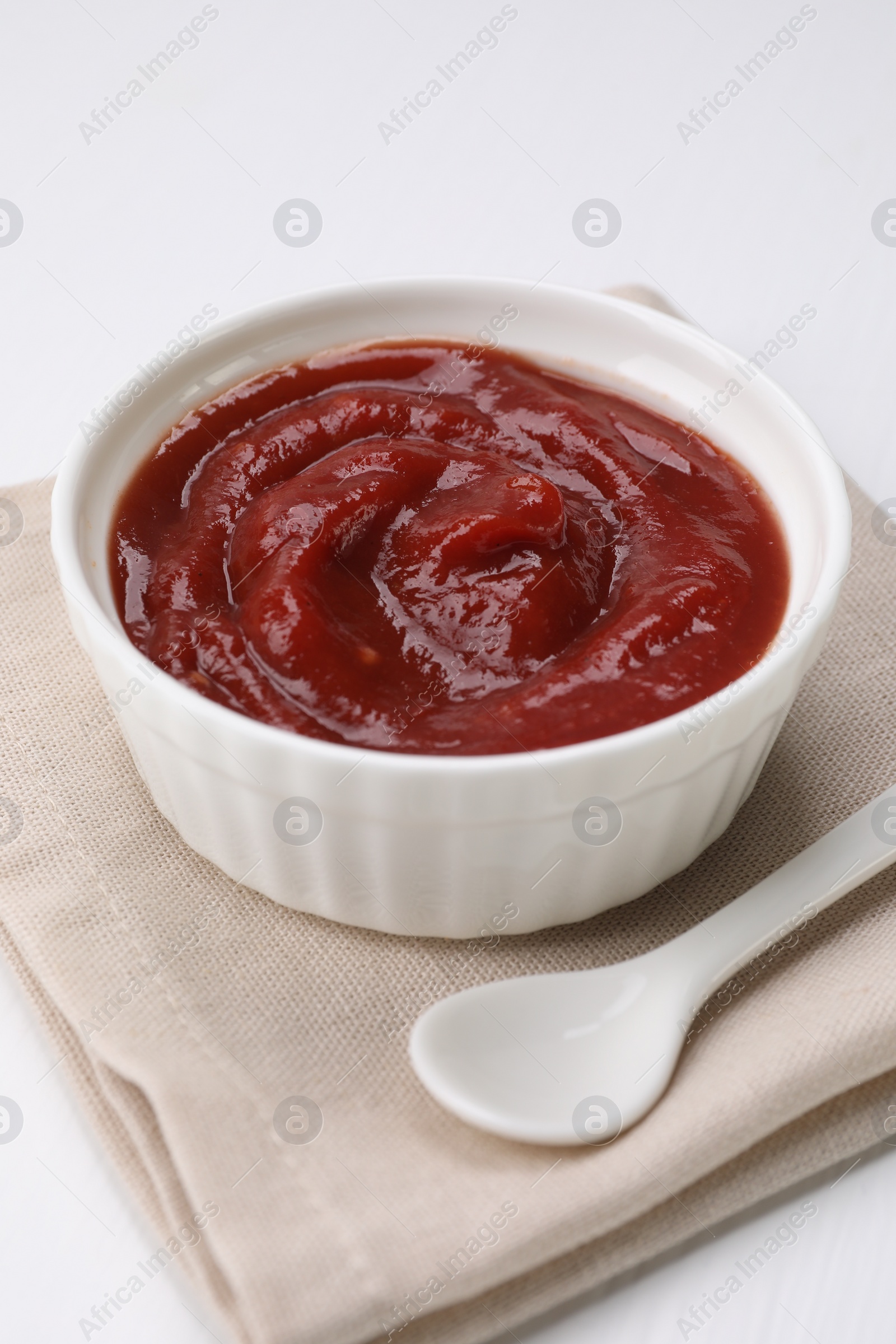 Photo of Organic ketchup in bowl and spoon on white table. Tomato sauce