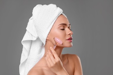 Young woman massaging her face with rose quartz roller on grey background
