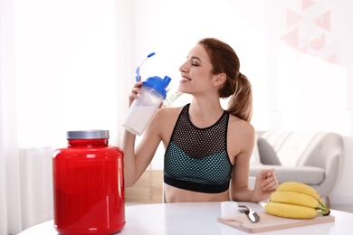 Young woman drinking protein shake at table with ingredients in room