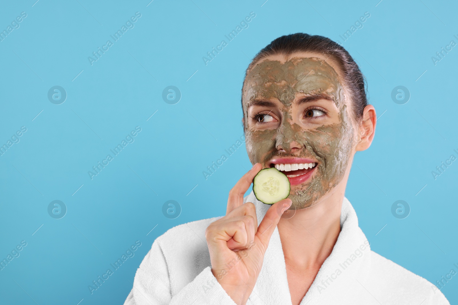 Photo of Beautiful woman with clay mask holding piece of cucumber on light blue background, space for text