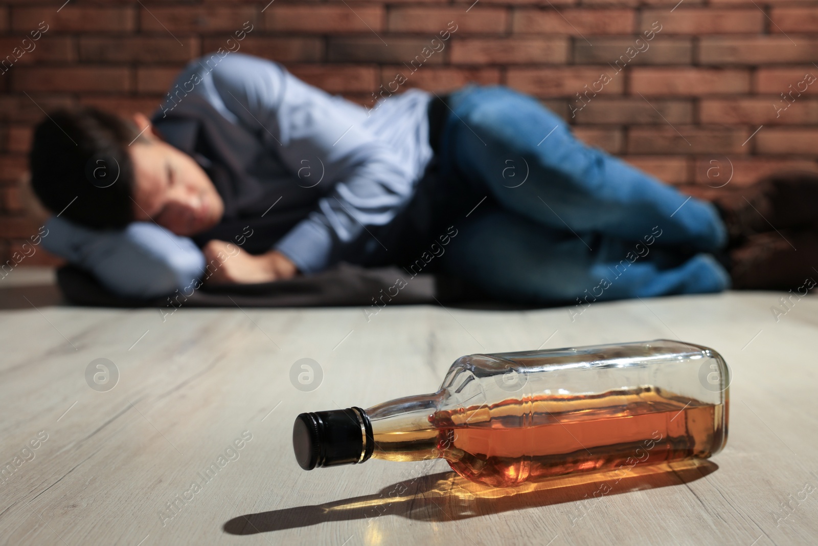 Photo of Addicted man lying near red brick wall, focus on bottle of alcoholic drink