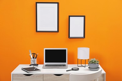 Photo of Workplace with laptop, stationery and magnifying glass on desk near orange wall. Home office