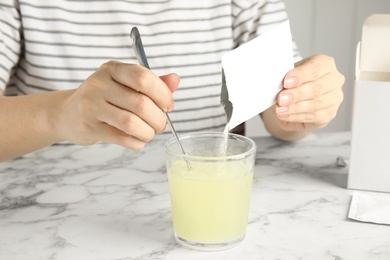 Woman pouring and stirring powder from medicine sachet into glass with water at table, closeup