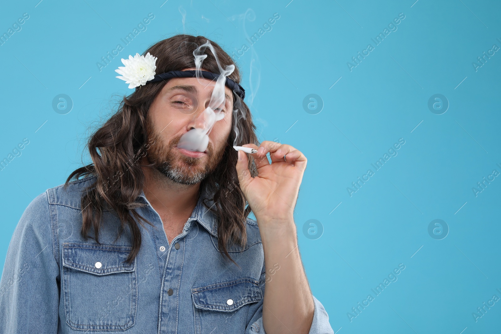 Photo of Stylish hippie man smoking cigarette on light blue background, space for text