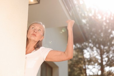 Photo of Angry senior woman showing fist near house, low angle view. Annoying neighbour