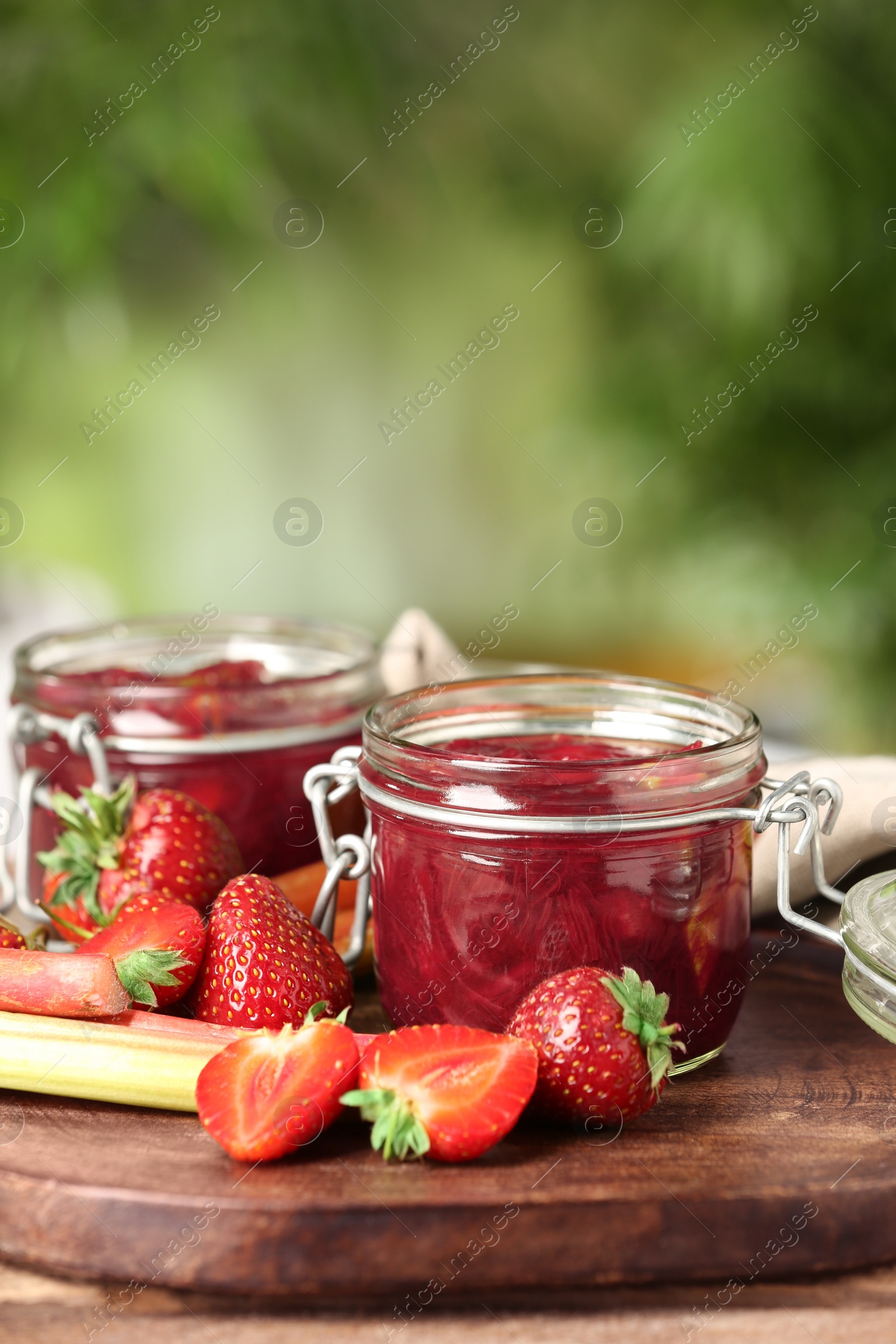 Photo of Tasty rhubarb jam in jars and strawberries on wooden table against blurred background. Space for text
