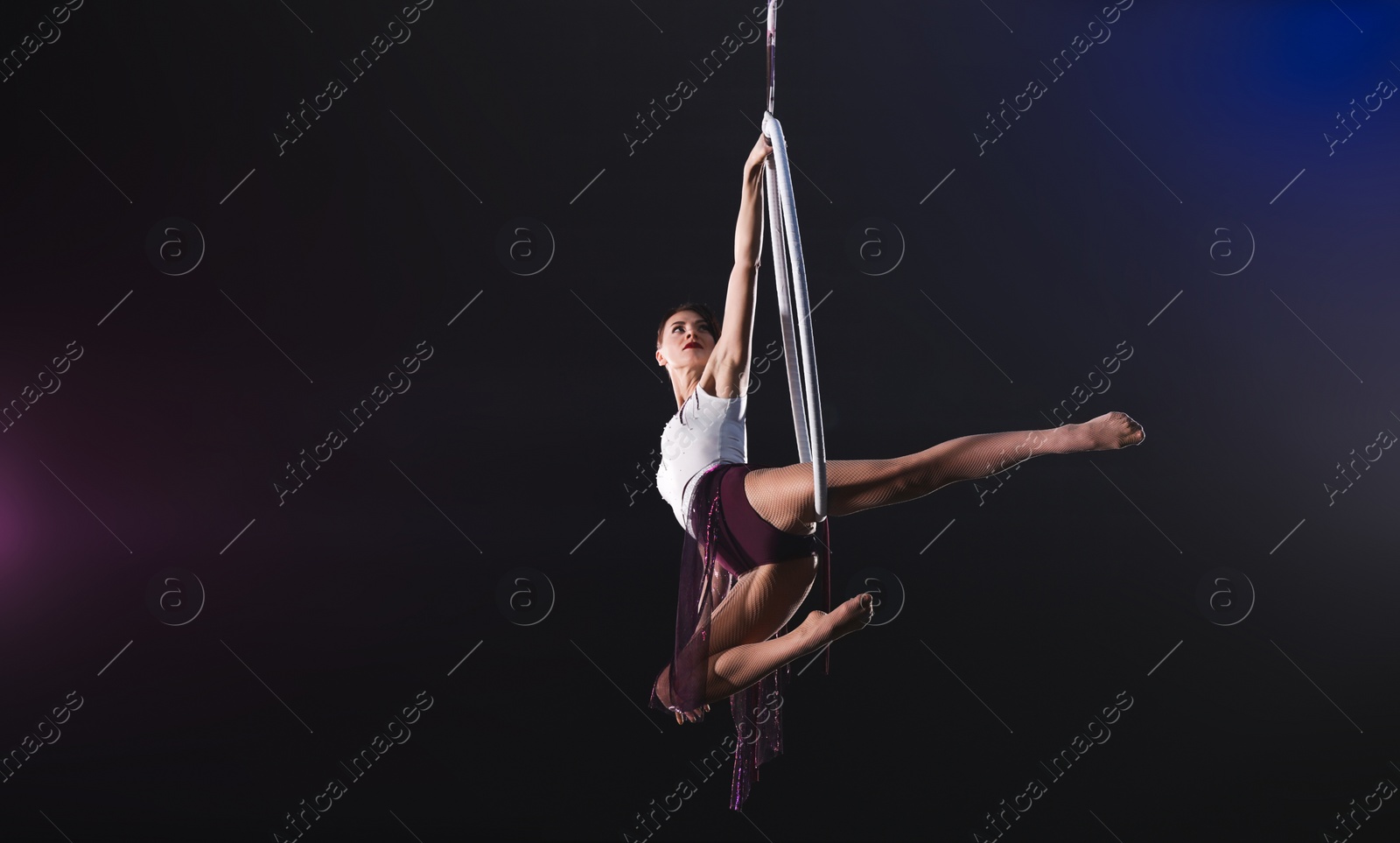 Photo of Young woman performing acrobatic element on aerial ring against dark background. Space for text