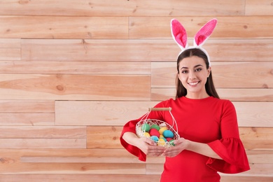 Photo of Beautiful woman in bunny ears headband holding basket with Easter eggs on wooden background, space for text
