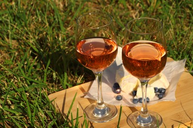 Photo of Wooden board with glasses of delicious rose wine, cheese and blueberries on green grass outdoors