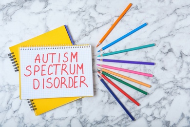 Photo of Notebook with words AUTISM SPECTRUM DISORDER and colorful pencils on marble background, top view
