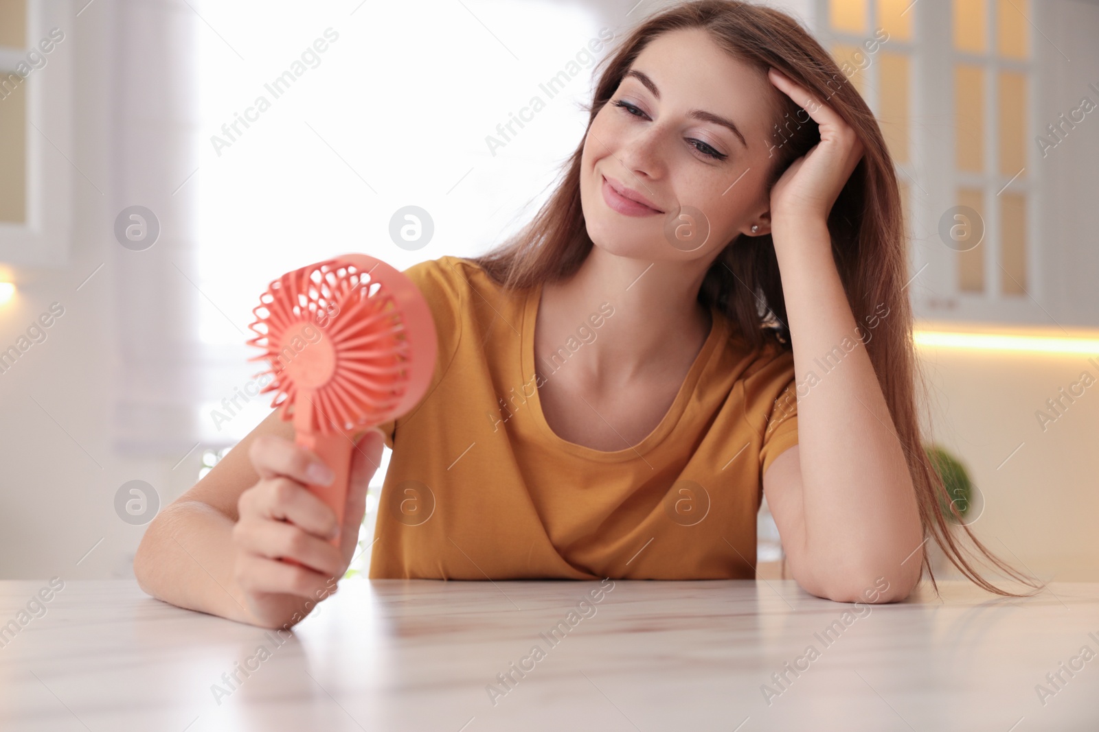 Photo of Woman enjoying air flow from portable fan at table in kitchen. Summer heat