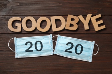 Photo of Text Goodbye 2020 and medical face masks on wooden background, flat lay