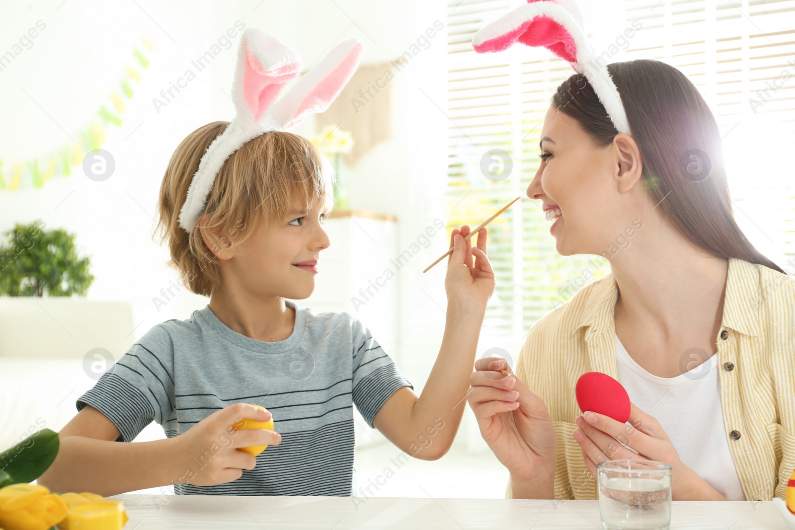 Photo of Happy mother and son with bunny ears headbands  having fun while painting Easter eggs at home