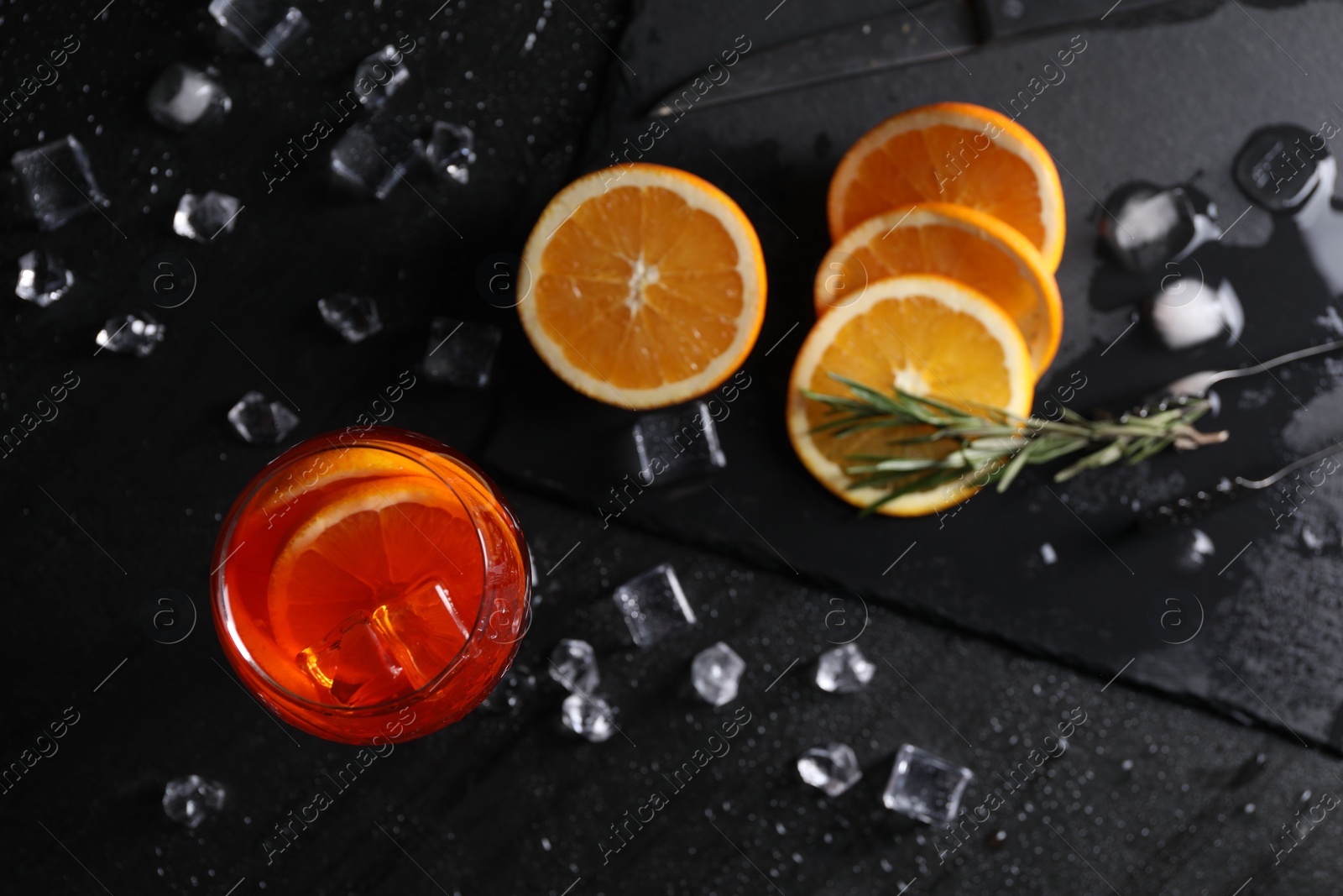 Photo of Glass of tasty Aperol spritz cocktail with orange slices and ice cubes on black table, flat lay