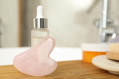 Photo of Rose quartz gua sha tool and cosmetic product on wooden bath caddy in bathroom, closeup. Space for text