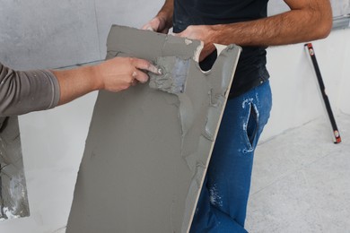 Photo of Workers applying cement on tile indoors, closeup