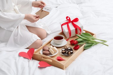 Photo of Tasty breakfast served in bed. Woman with I Love You card, desserts, tea, flowers and gift box at home, closeup