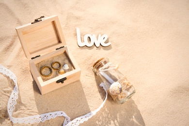 Photo of Wooden box with gold wedding rings, invitation in glass bottle and word Love on sandy beach