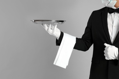 Photo of Waiter in medical face mask holding tray on light grey background, closeup