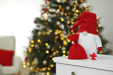 Photo of Funny Christmas gnome with bag on commode in room with festive decoration. Space for text