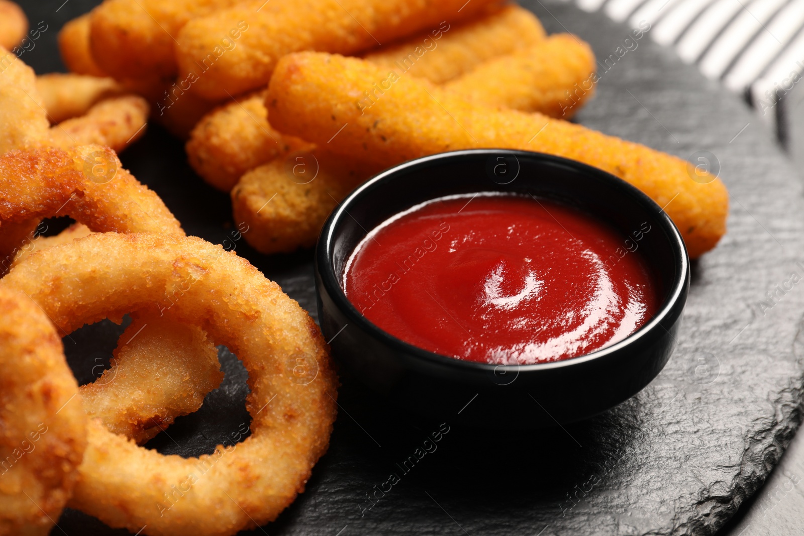 Photo of Tasty ketchup, onion rings and cheese sticks on black slate board, closeup