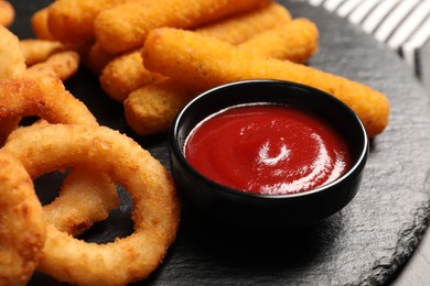Photo of Tasty ketchup, onion rings and cheese sticks on black slate board, closeup