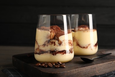 Photo of Delicious tiramisu in glasses, spoon and coffee beans on wooden board, closeup