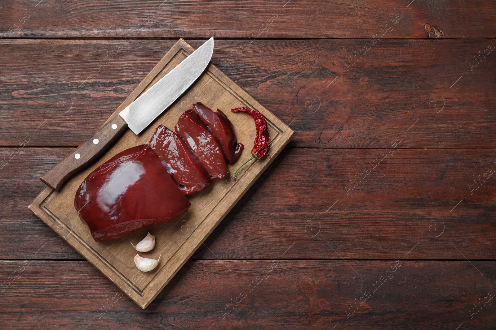 Photo of Cut raw beef liver with chili pepper, garlic and knife on wooden table, top view. Space for text