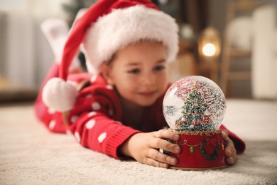 Little girl in Santa hat playing with snow globe on floor