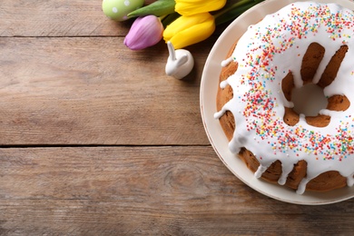 Photo of Glazed Easter cake with sprinkles, decorative bunny, painted egg and tulips on wooden table, flat lay. Space for text