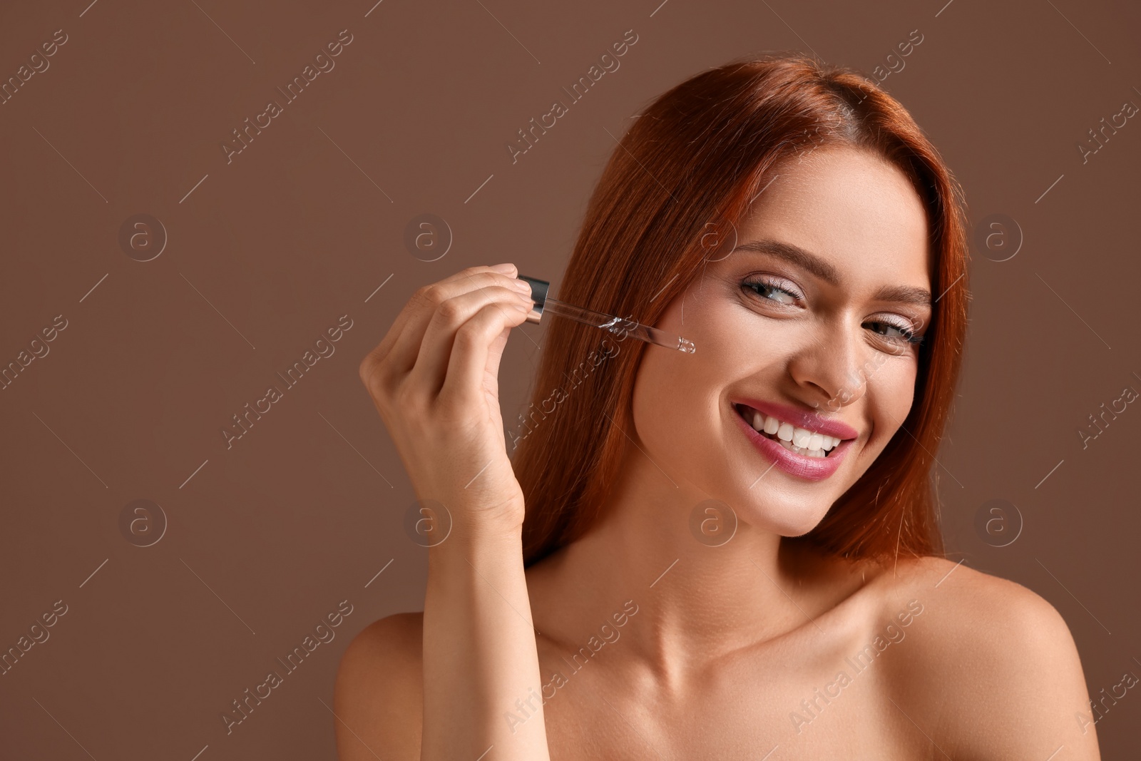 Photo of Beautiful young woman applying cosmetic serum onto her face on brown background, space for text