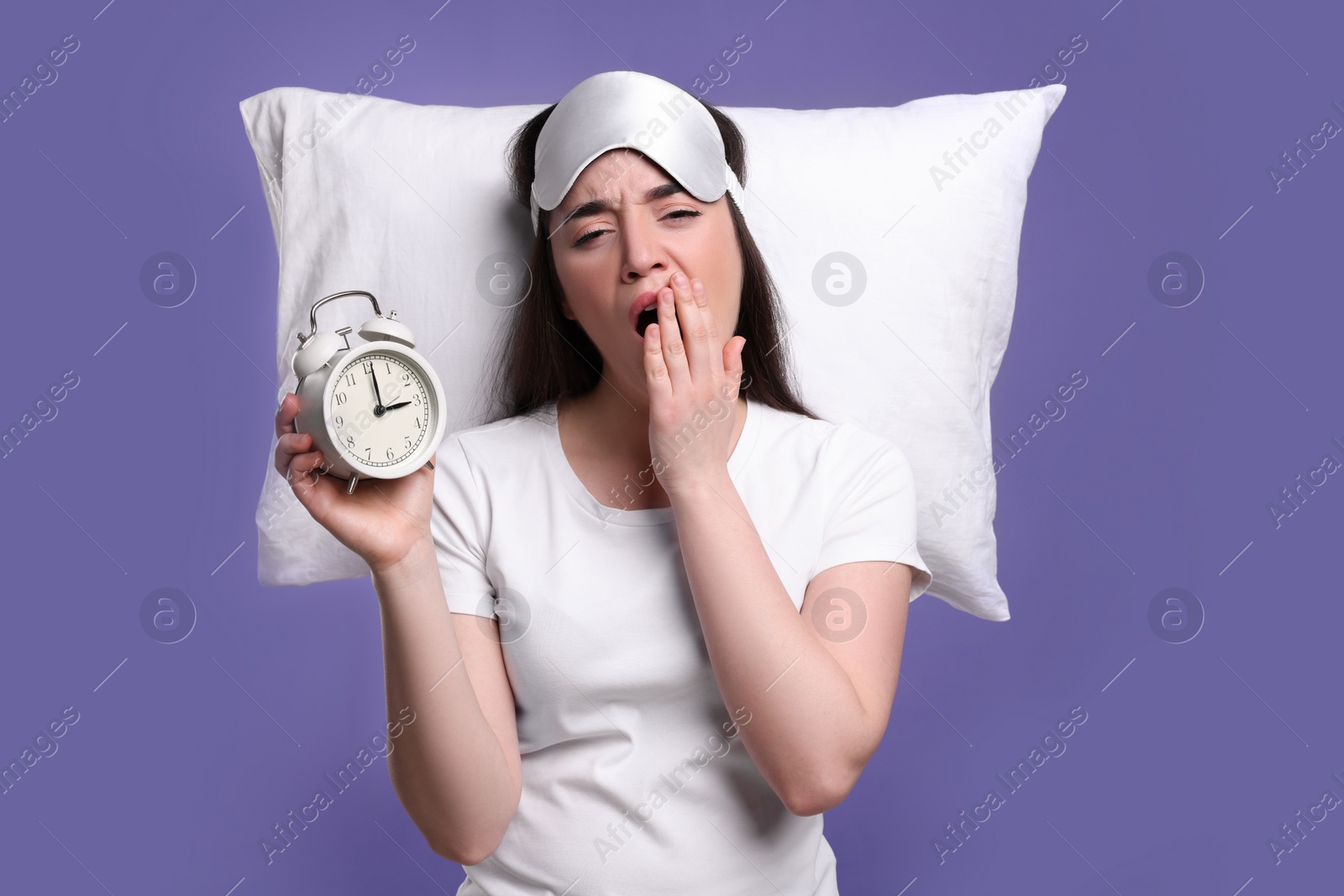 Photo of Tired young woman with sleep mask, alarm clock and pillow on purple background. Insomnia problem