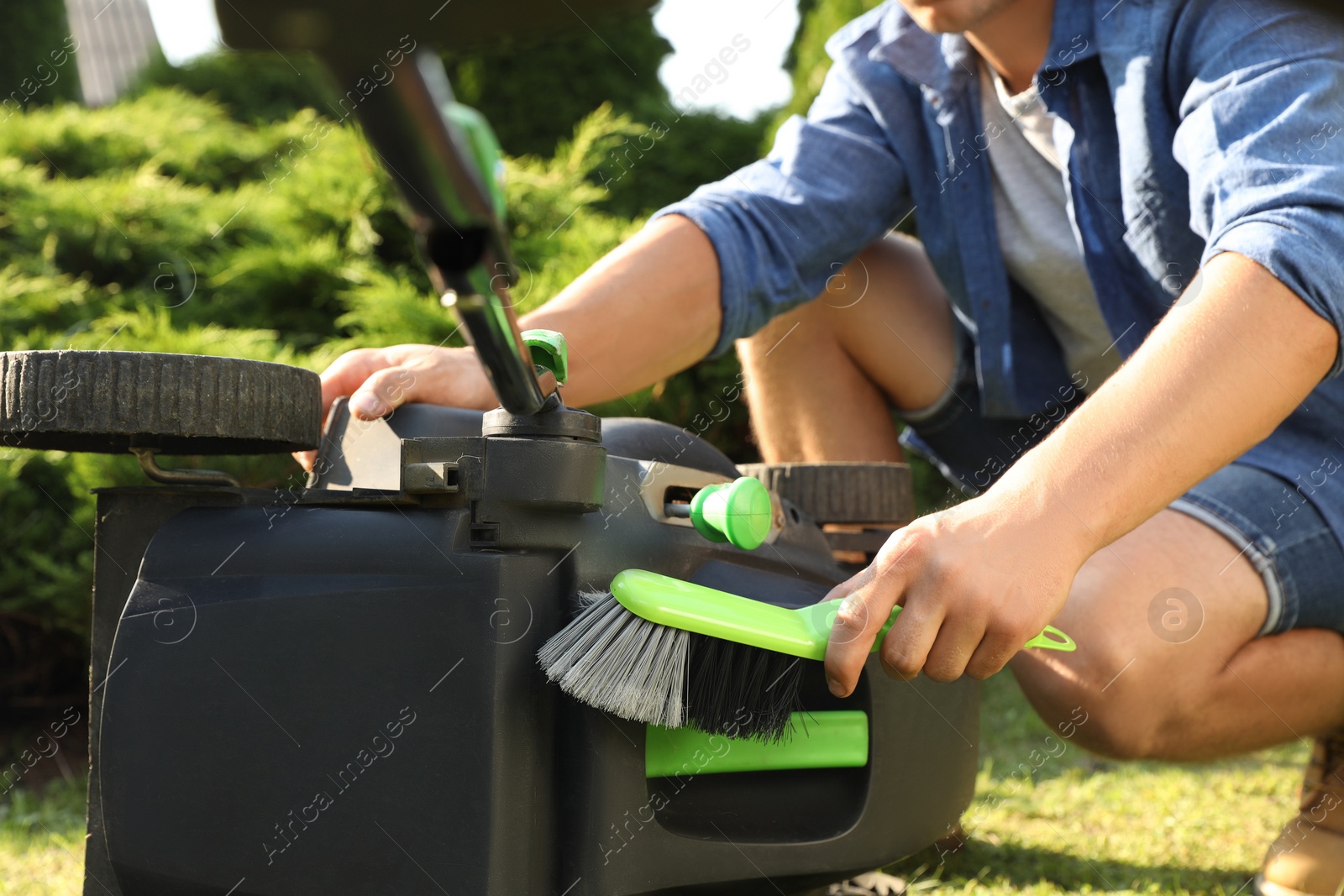 Photo of Young man cleaning lawn mower with brush in garden, closeup