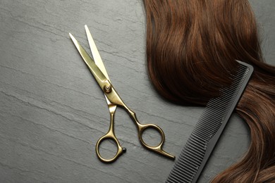 Photo of Professional hairdresser scissors and comb with brown hair strand on dark grey table, top view