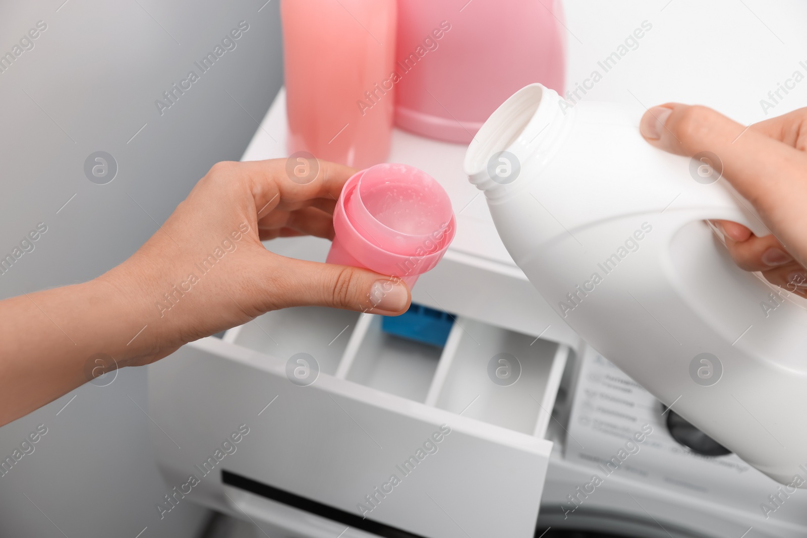 Photo of Woman pouring laundry detergent from bottle into cap near washing machine, closeup