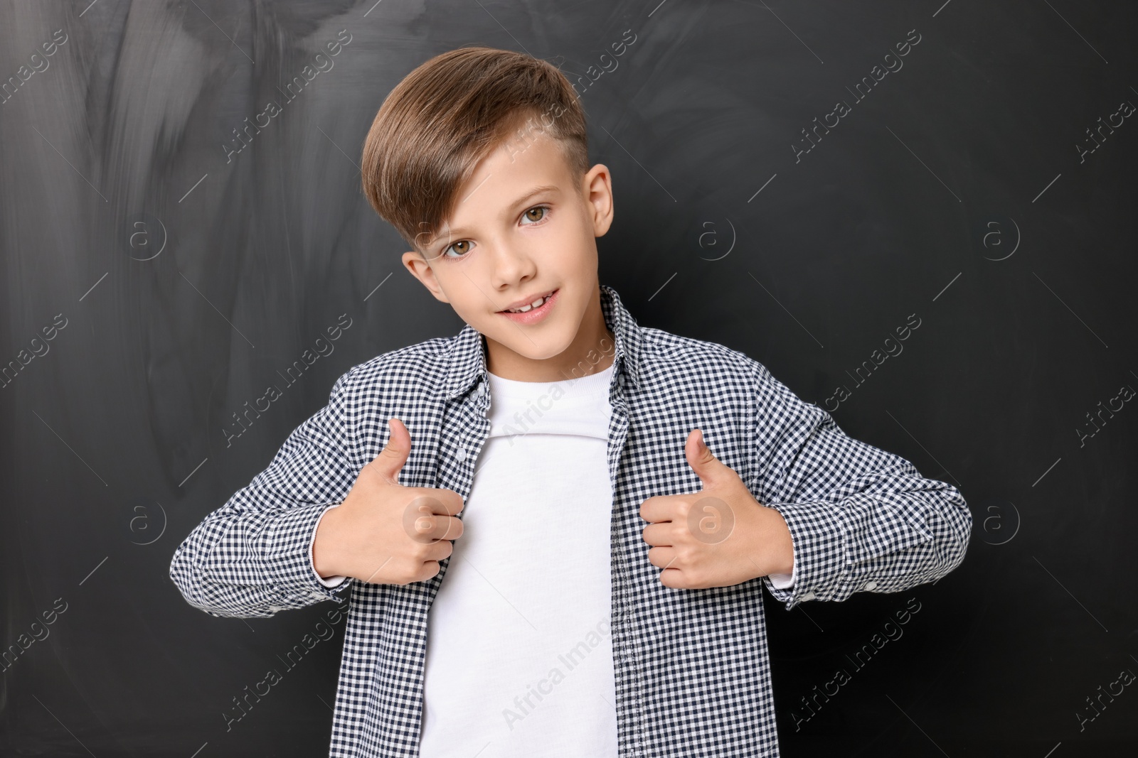 Photo of Cute schoolboy showing thumbs up near chalkboard