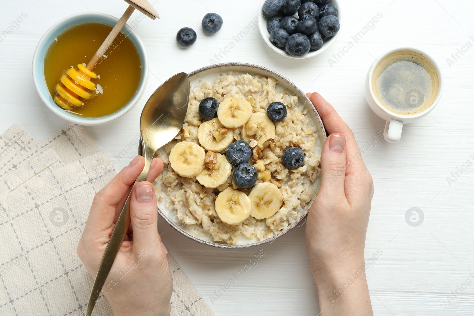 Photo of Woman eating tasty oatmeal with banana, blueberries, walnuts and honey at white wooden table, top view