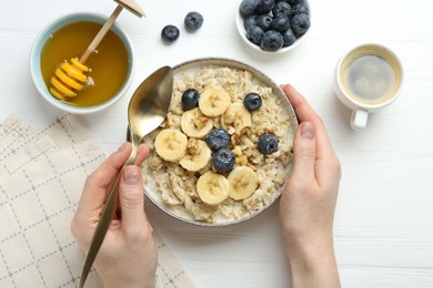 Woman eating tasty oatmeal with banana, blueberries, walnuts and honey at white wooden table, top view